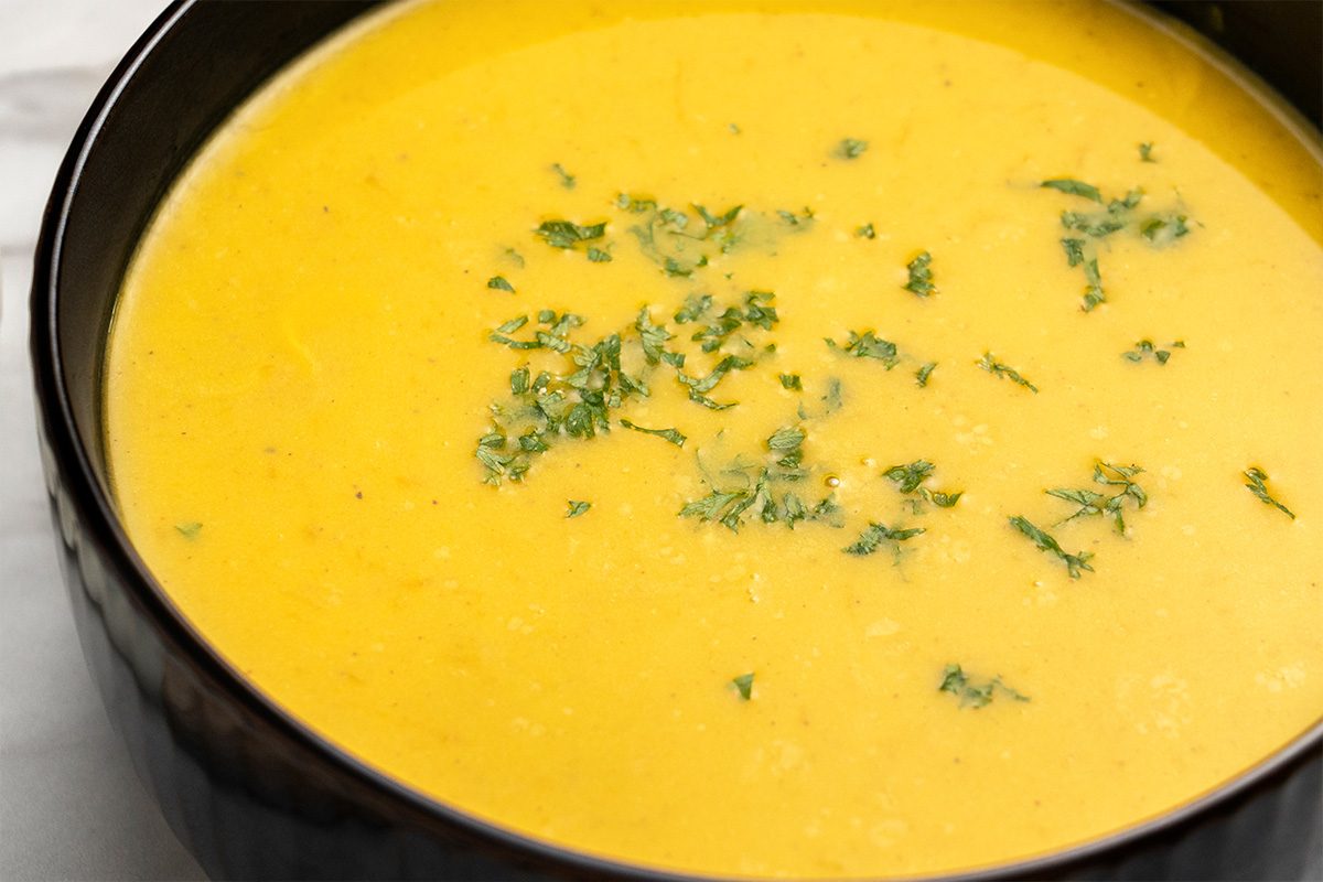 A bowl of creamy yellow soup garnished with chopped fresh herbs, served in a dark-colored bowl on a light surface.