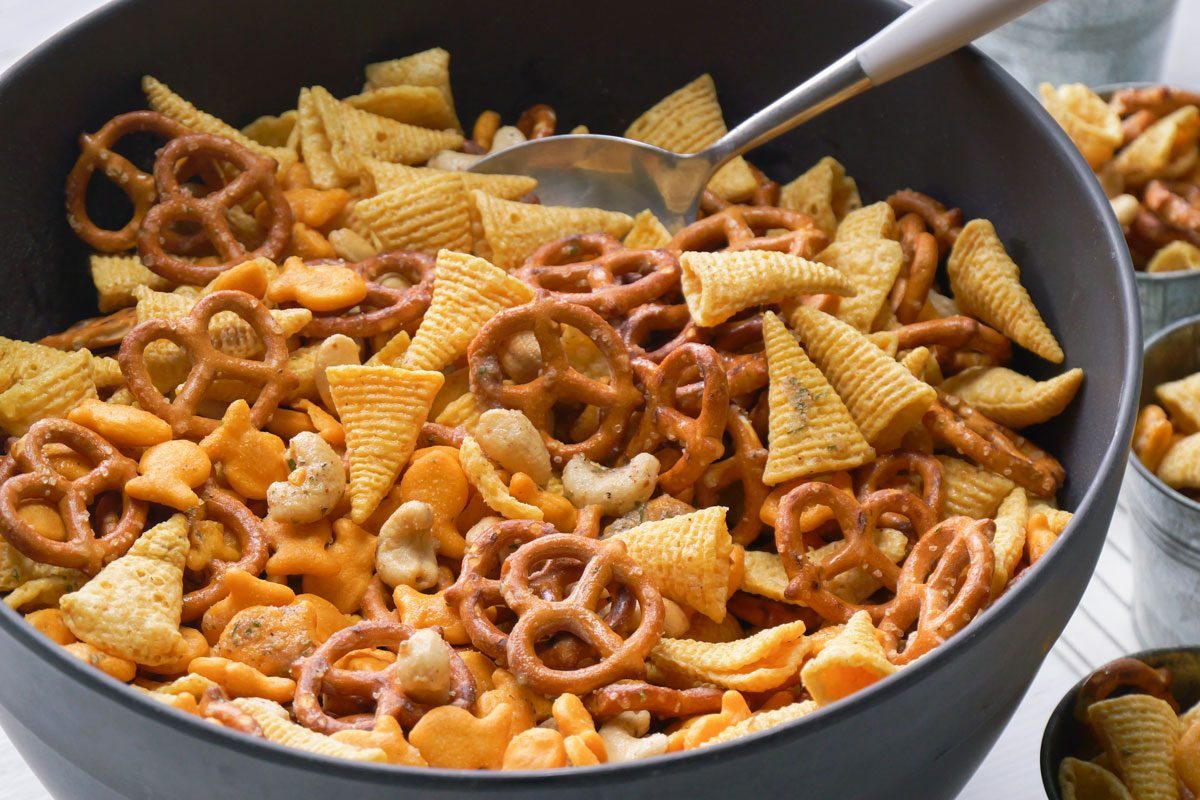A large black bowl holds a colorful snack mix featuring pretzels; cheese crackers; corn chips; and nuts. A metal serving spoon is inside; with extra bowls of the snack mix seen in the blurred background