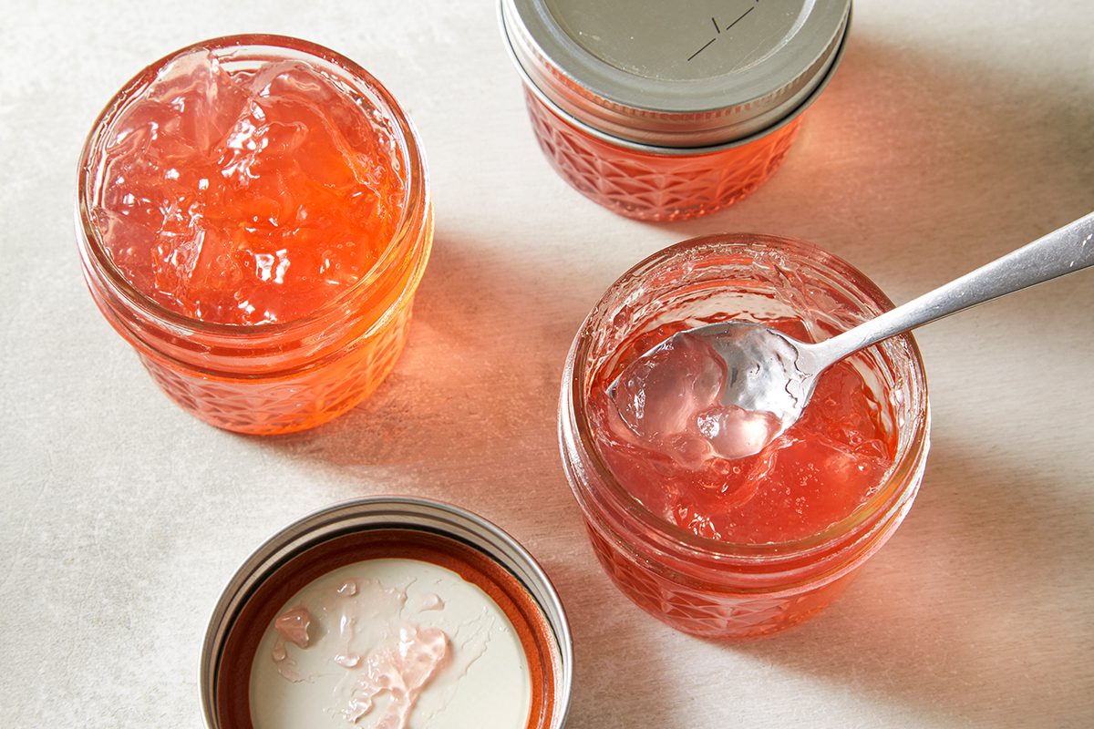 Three small glass jars filled with orange jelly sit on a light surface. One jar is open with a spoon inside, and two lids are nearby, one resting on top of a jar.