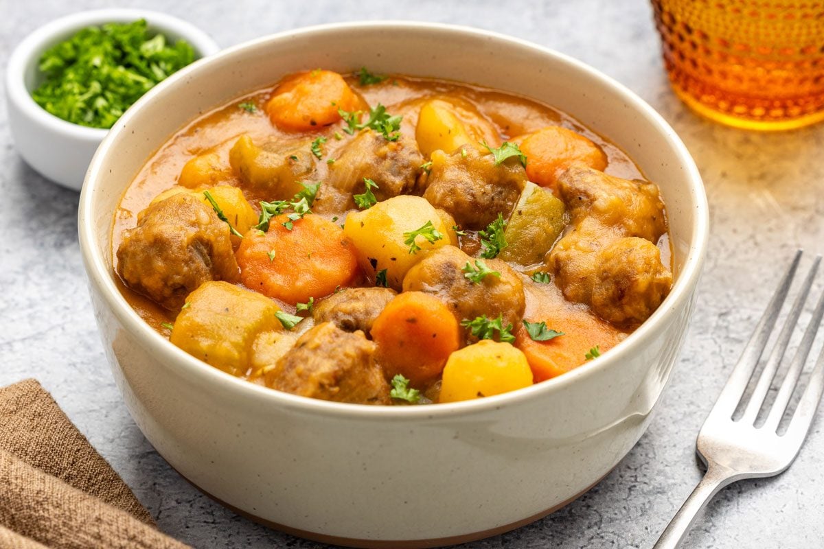 Close shot of Homemade Italian Sausage Stew in a white bowl with a drink; garinished with cilantro; a fork and a napkin nearby; all set on grey marble surface;