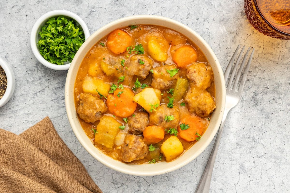 Overhead shot of Homemade Italian Sausage Stew in a white bowl with a drink; garinished with cilantro; a fork and a napkin nearby; all set on grey marble surface;