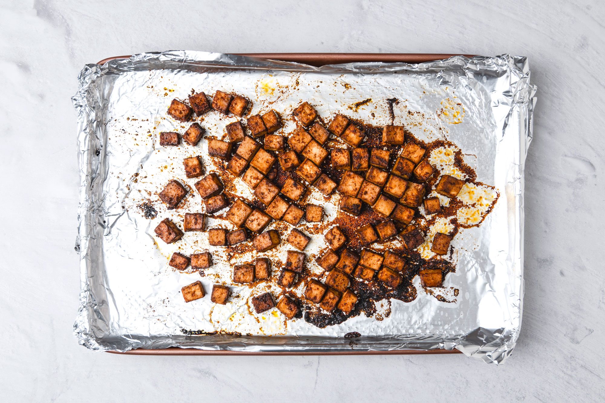 A top shot of Golden-brown roasted tofu cubes; coated with seasoning; arranged on a baking sheet lined with aluminum foil; set atop a light marble surface