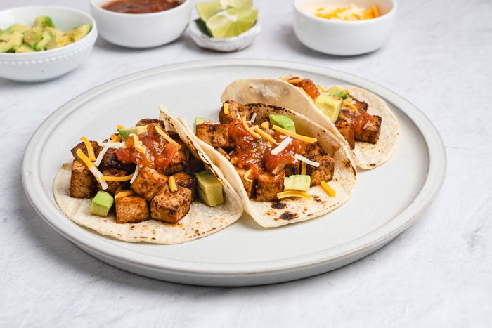 3/4th shot of Three soft tacos filled with seasoned tofu cubes, diced avocado, shredded cheese, and salsa are served on a plate, In the background, small bowls hold extra avocado, shredded cheese, lime wedges, and salsa