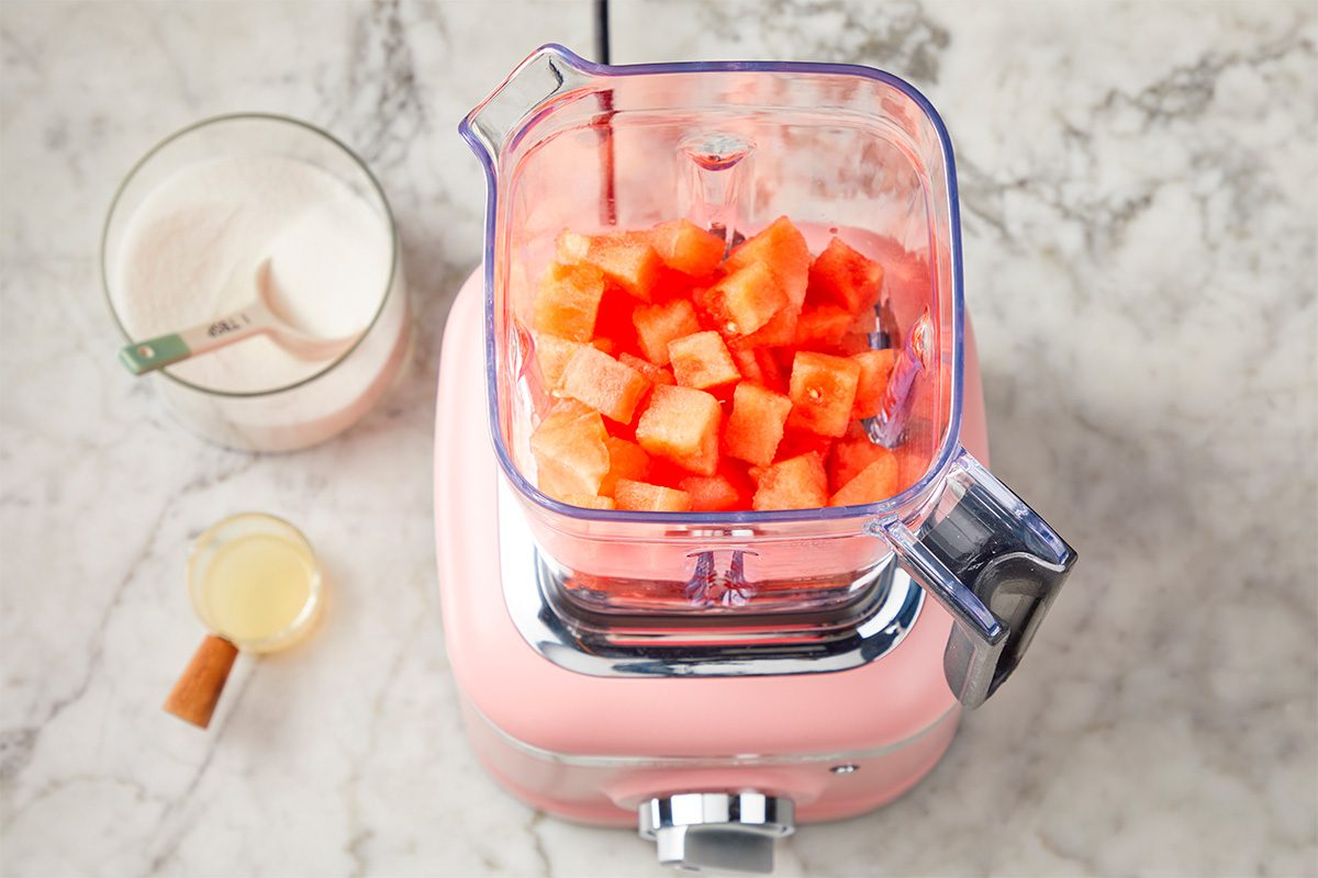 A pink blender filled with cubed watermelon sits on a marble countertop. Nearby are a measuring cup of sugar, a small glass of liquid, and a cinnamon stick.