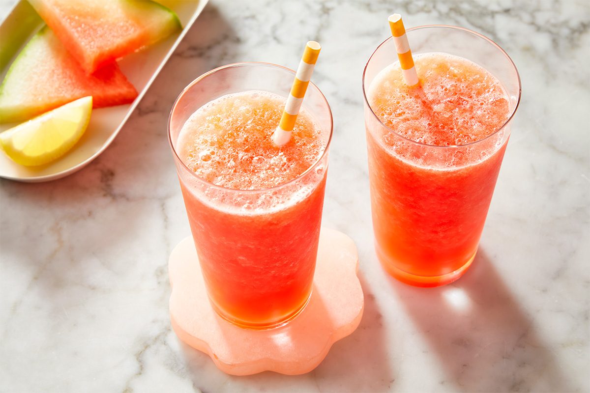 Two tall glasses of watermelon juice with striped straws sit on a marble surface, beside a plate with watermelon slices and a lemon wedge.