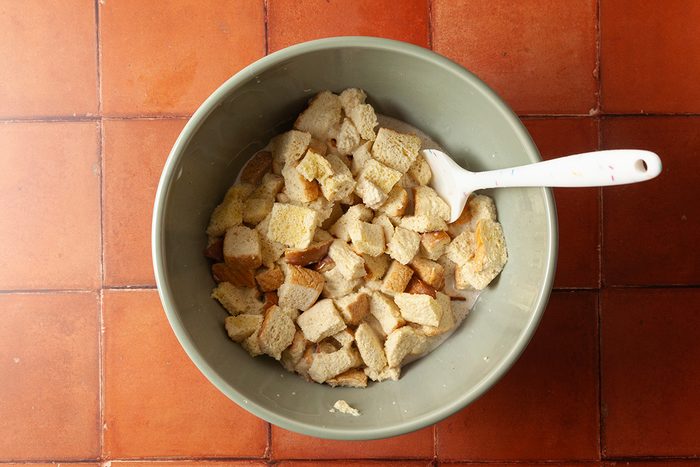 A green bowl filled with chunks of bread soaking in liquid, with a white spoon resting inside, on a red tiled surface.