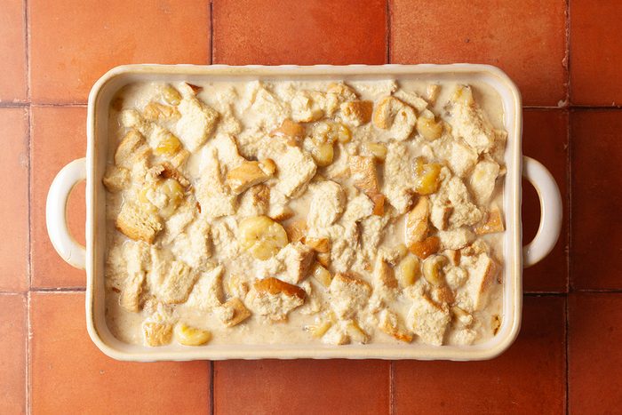 A rectangular baking dish filled with unbaked bread pudding mixture, showing chunks of bread and bananas in a creamy liquid, placed on a tiled surface.
