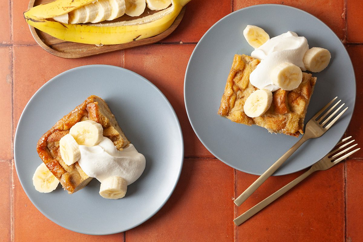 Two blue plates each with a slice of banana-topped pastry, whipped cream, and banana slices. A peeled banana on a wooden tray is in the background. Two gold forks are on the right plate.