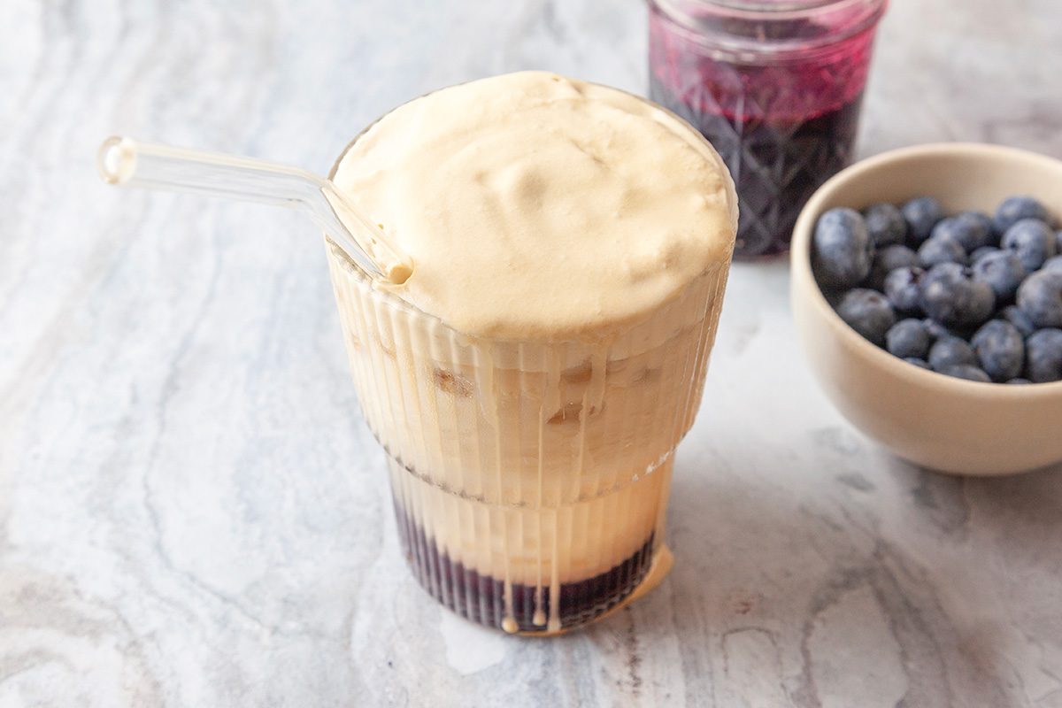 Close-up shot of a layered iced drink with a frothy cream top in a ribbed glass with a glass straw; set on a light marbled surface; a bowl of blueberries and a jar of blueberry syrup are in the background