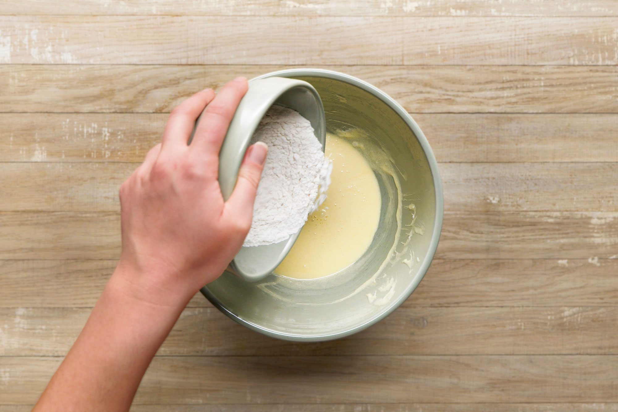 overhead shot of a hand pours flour from a small bowl into a larger bowl containing a yellow batter, on a light wooden surface