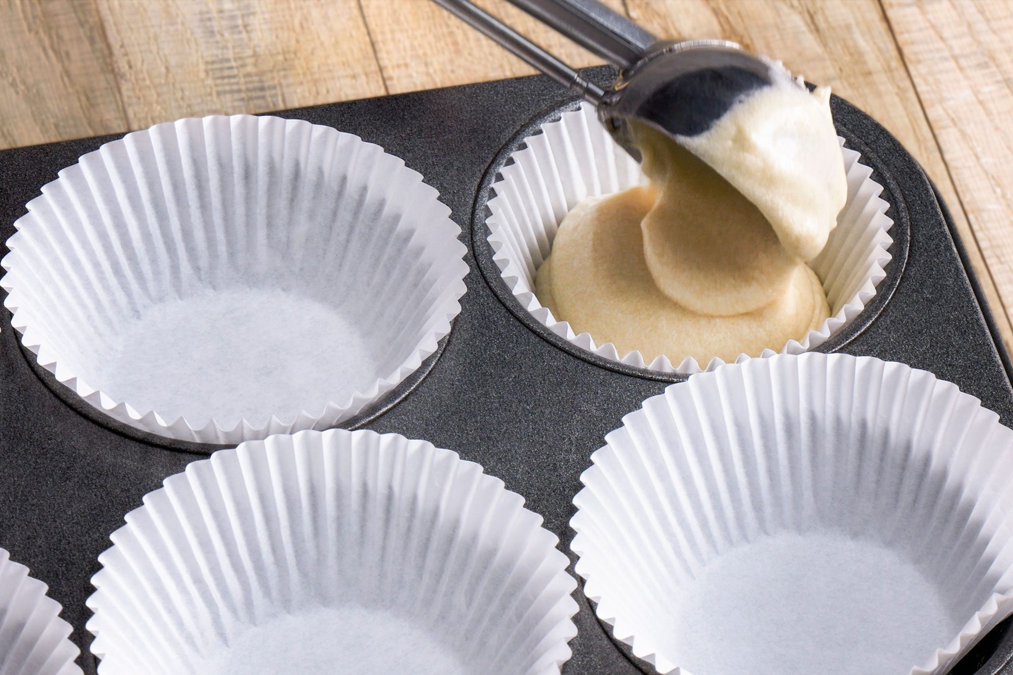 3/4th shot of a metal ice cream scoop fills a white paper cupcake liner with batter in a muffin tin, while other empty liners wait to be filled on a wooden surface