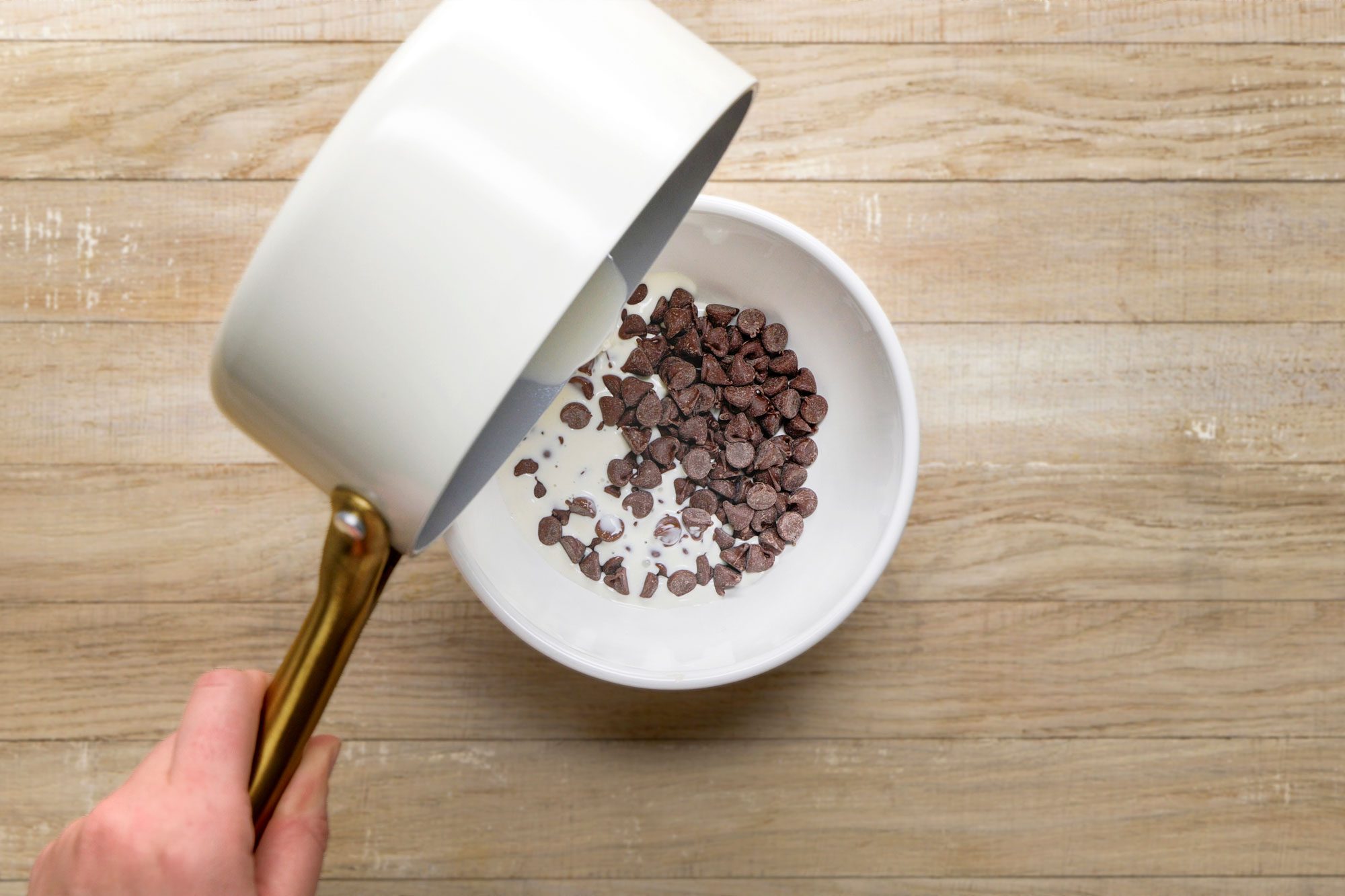 overhead shot of a hand pours liquid from a white saucepan into a white bowl filled with chocolate chips, on a light wooden surface