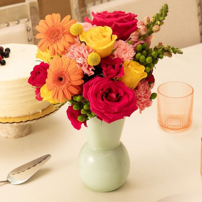A pastel green vase filled with vibrant flowers including red roses, yellow and orange blooms, sits on a white table next to a cake, a glass, and a serving utensil.