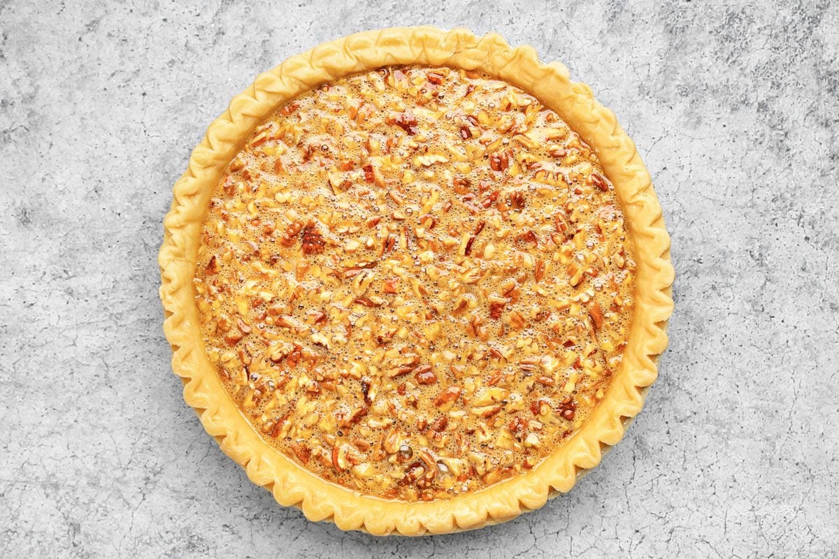 overhead shot of a whole pecan pie with a crimped golden brown crust, filled with a shiny, nutty pecan filling, sits on a light gray textured surface