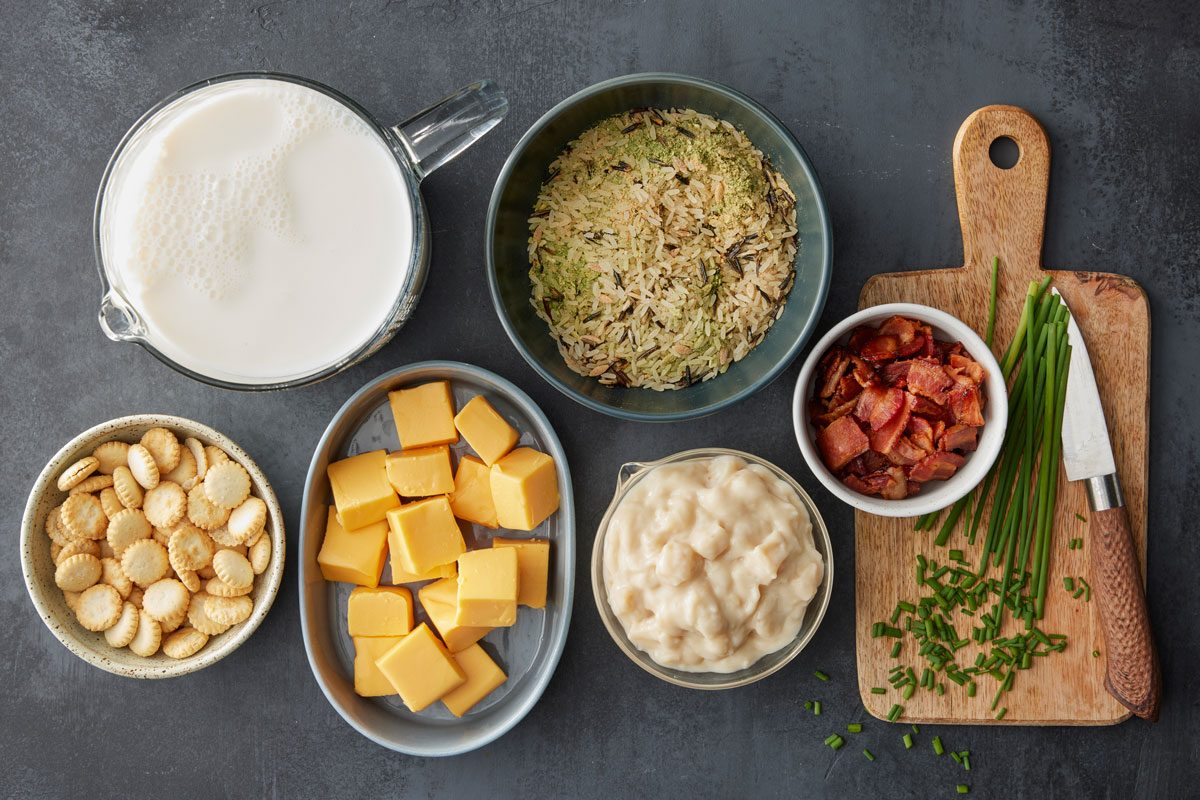 overhead shot of a milk in a glass pitcher, wild rice in a bowl, chopped cooked bacon, oyster crackers, cubed cheddar cheese, creamy soup, and fresh chives with a knife on a small cutting board