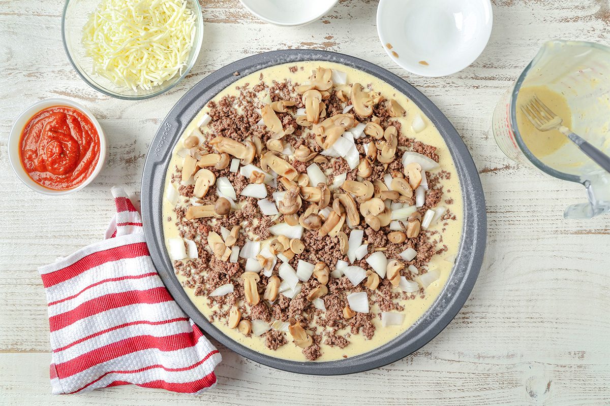 A pizza crust topped with ground beef, mushrooms, and chopped onions sits on a round pan. Surrounding it are bowls of shredded cheese, tomato sauce, and a striped red and white towel on a white wooden surface.
