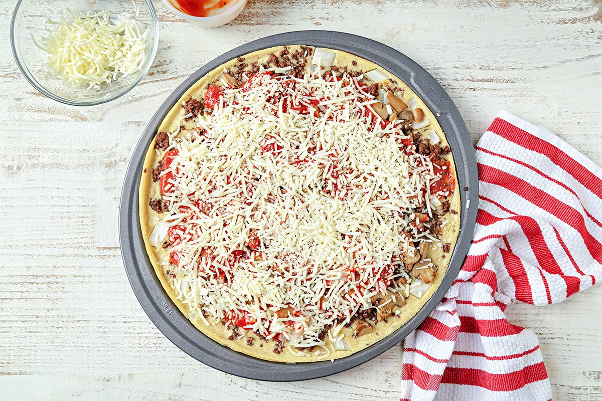 A pizza topped with ground meat, diced tomatoes, mushrooms, and shredded cheese sits on a round baking pan. A bowl of cheese and a red-and-white striped towel are nearby on a light wooden surface.