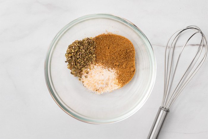 A glass bowl with three different spices or seasonings, separated but touching, next to a metal whisk on a white marble surface.