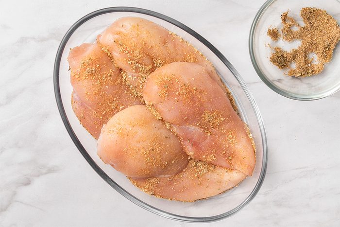 Four raw chicken breasts in a glass baking dish, sprinkled with a dry seasoning mix. A bowl with extra seasoning mix sits nearby on a white marble surface.