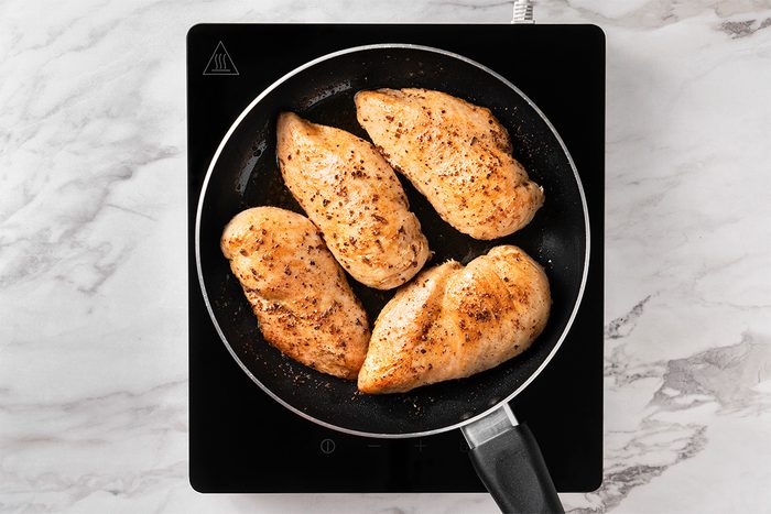 Four seasoned chicken breasts are cooking in a black frying pan on an electric stovetop, set on a white marble countertop. The chicken is lightly browned and arranged to fit within the pan.