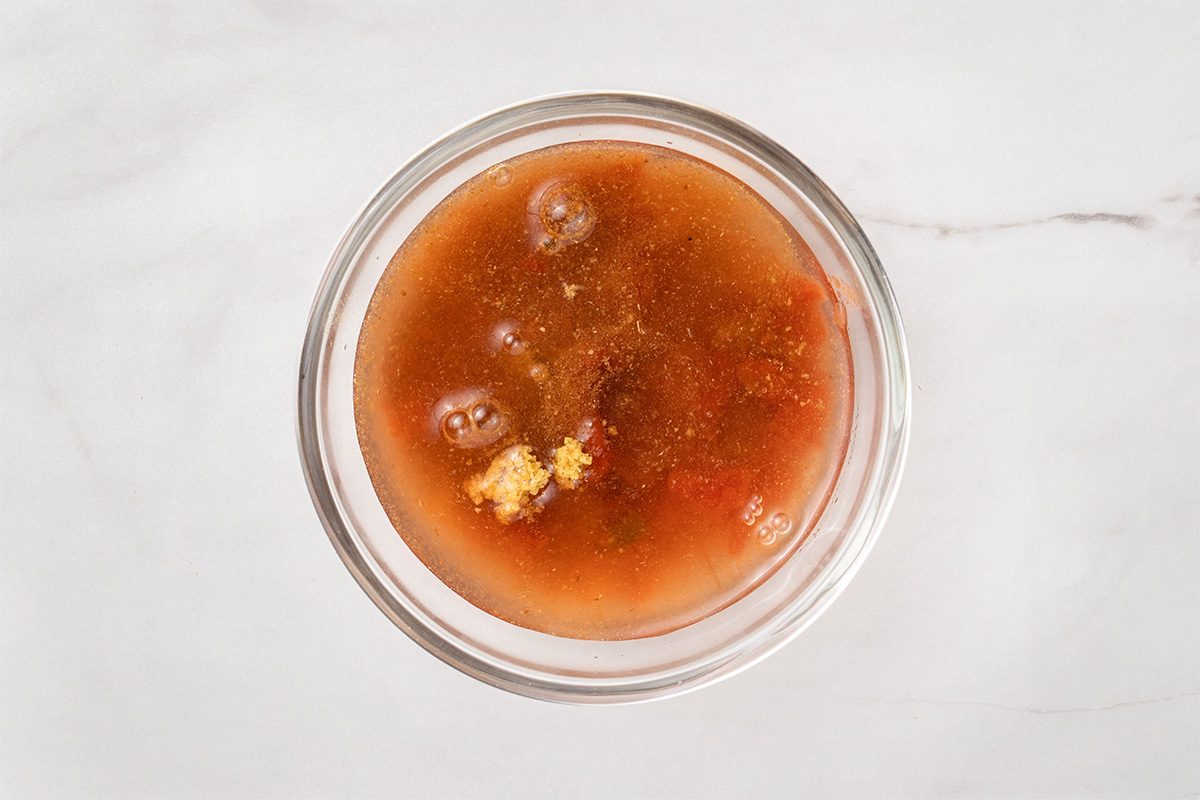 A clear glass bowl filled with a reddish-brown liquid soup containing visible chunks of vegetables and seasonings, placed on a white marble surface.