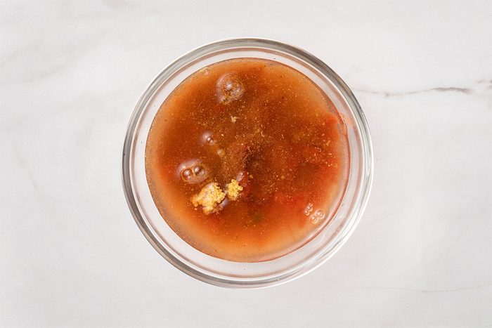 A clear glass bowl filled with a reddish-brown liquid soup containing visible chunks of vegetables and seasonings, placed on a white marble surface.
