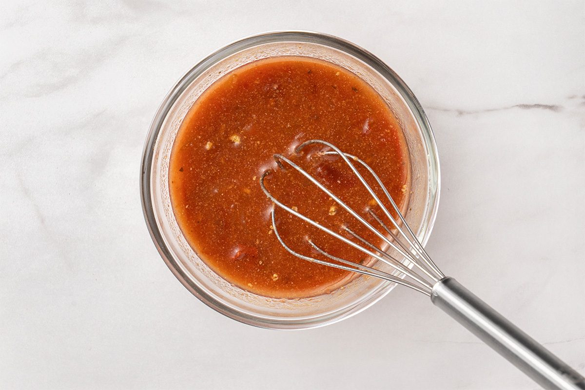 A metal whisk in a glass bowl filled with reddish-brown sauce or marinade, placed on a light marble surface.
