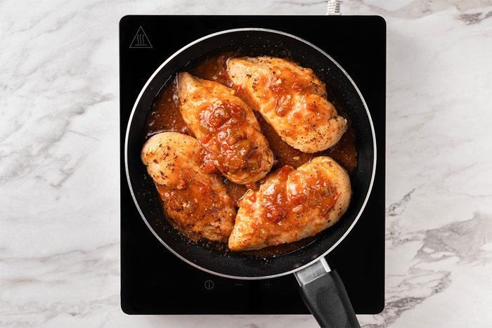Four seasoned chicken breasts cooking in a frying pan on a black induction cooktop, topped with a chunky tomato-based sauce. The background is a white marble countertop.