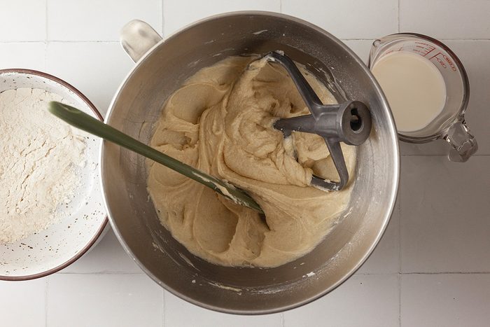 A mixing bowl with cookie dough, a green spatula, and a mixer attachment sits between a bowl of flour and a measuring cup of milk on a white surface.