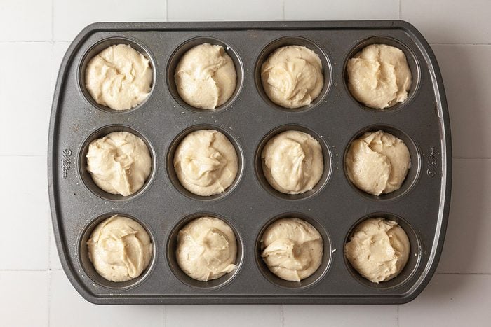 A muffin tin filled with twelve portions of unbaked muffin batter, ready to be baked, sits on a white tiled surface.
