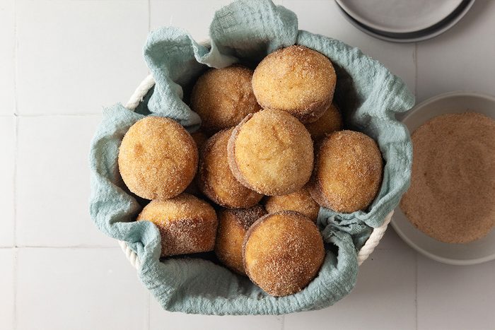 A basket lined with a light blue cloth holds a dozen golden brown, sugar-coated doughnuts. A plate with cinnamon sugar is partially visible beside the basket, all placed on a white tiled surface.