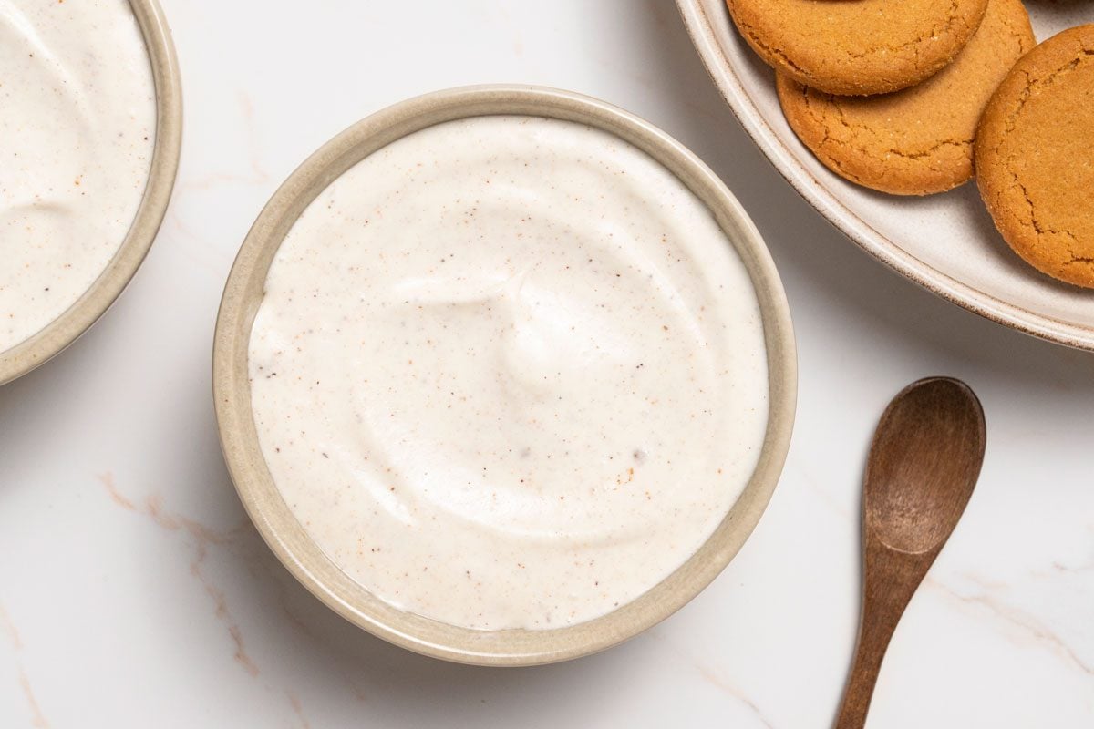 Overhead shot of Gingersnap Dip served in bowsl; a wooden spoon and a plate of gingersnaps cookies are nearby visible; all set on a white marble surface;