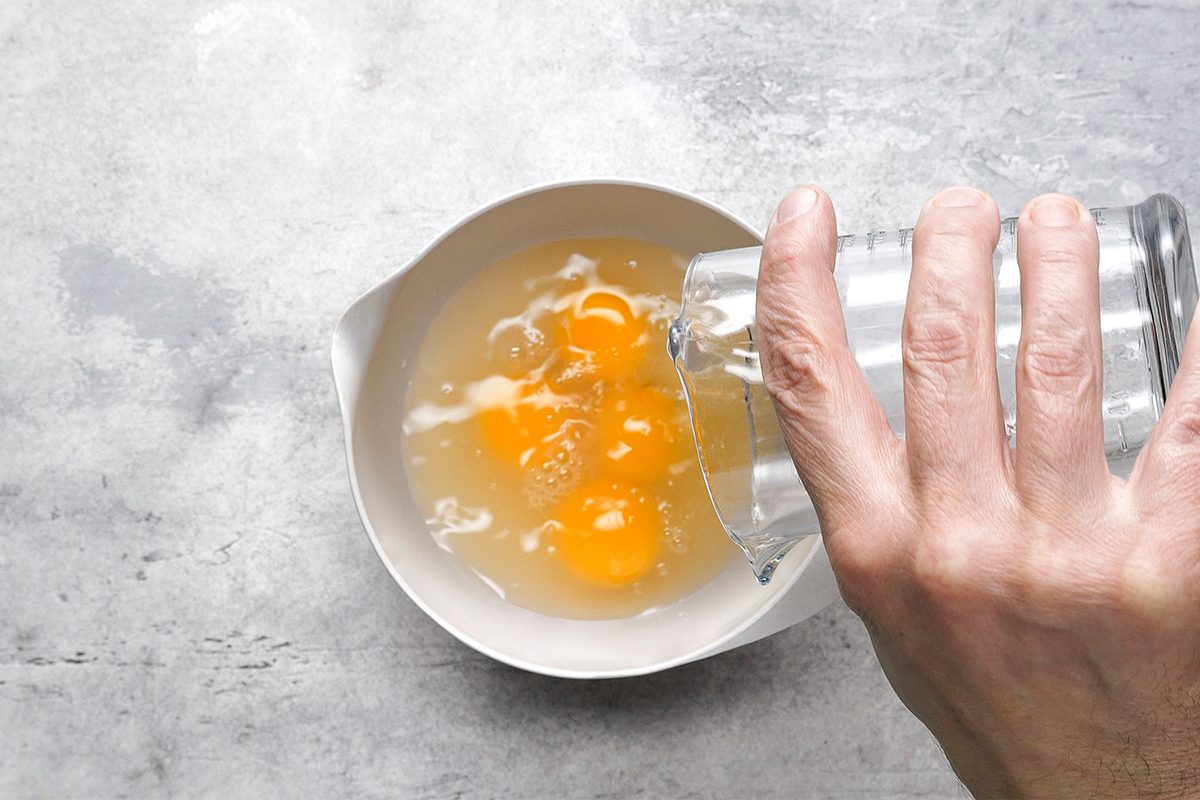 A hand pours water from a glass into a white bowl containing cracked eggs, set on a gray countertop.
