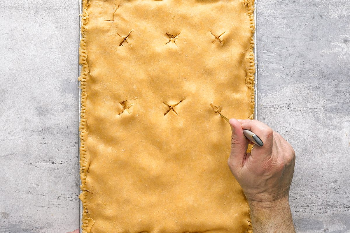 A hand uses a fork to poke holes in the top of a large, rectangular unbaked pie crust on a baking sheet. The crust has several vent slits already cut. The background is a gray countertop.