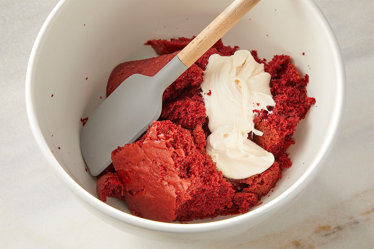 A white bowl contains crumbled red velvet cake and a dollop of cream cheese frosting. A spatula with a wooden handle rests inside the bowl, ready to mix the ingredients together.