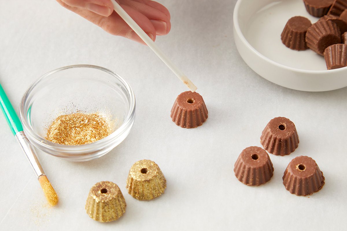 A hand uses a tool to apply edible gold glitter to small, ridged chocolate candies. A brush and a bowl of gold glitter are nearby, with more chocolates on a white plate in the background.