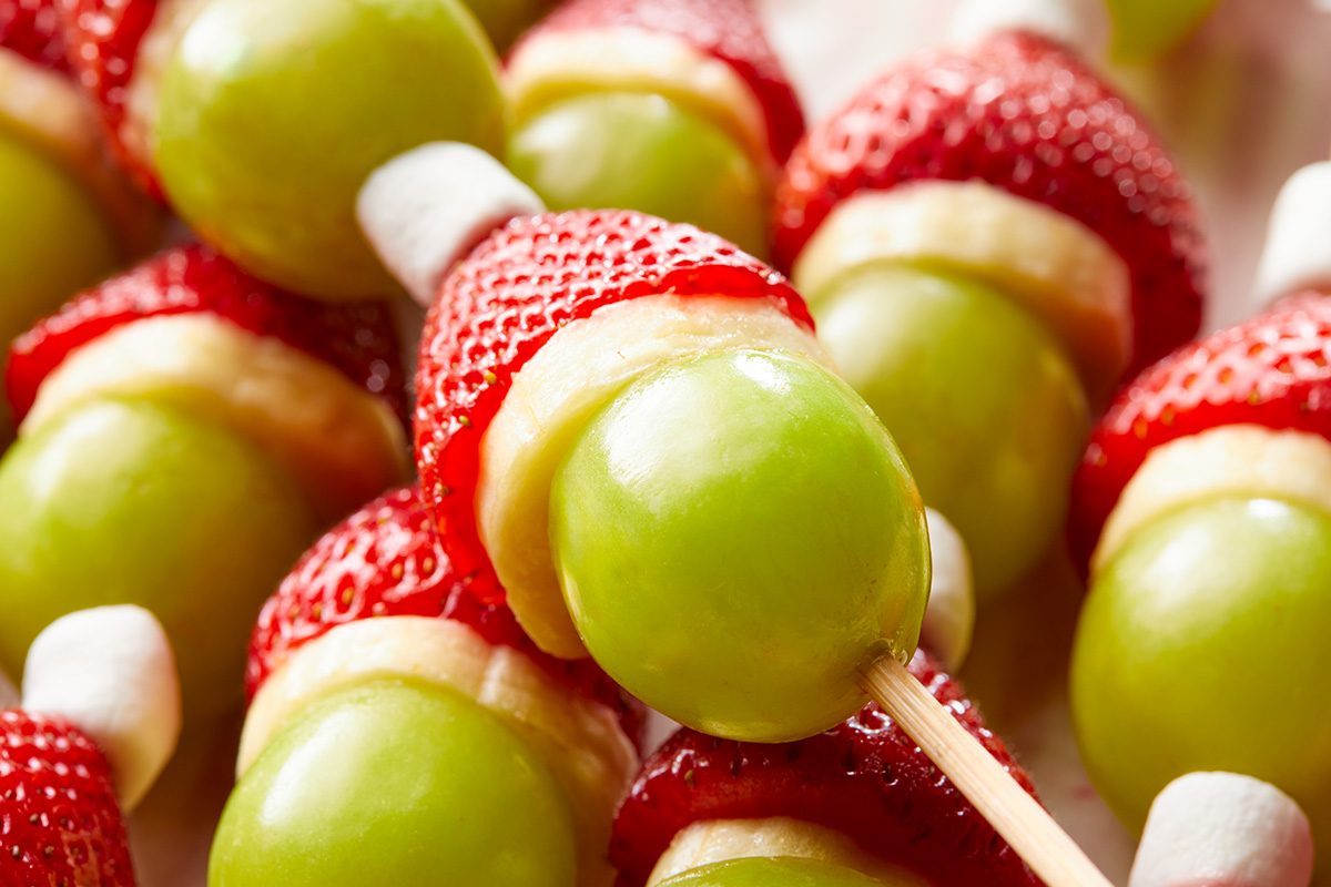 Close up shot of fruit skewers with green grapes, banana slices, strawberries, and mini marshmallows assembled to look like festive holiday treats, resembling Christmas themed snacks