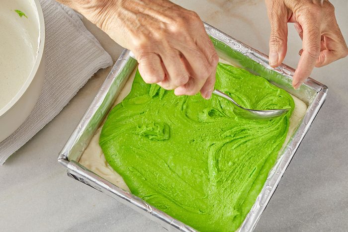 Overhead shot of a person uses a spoon to spread a layer of bright green batter over a white layer in a foil lined square baking pan; with a bowl and towel nearby on a marble surface