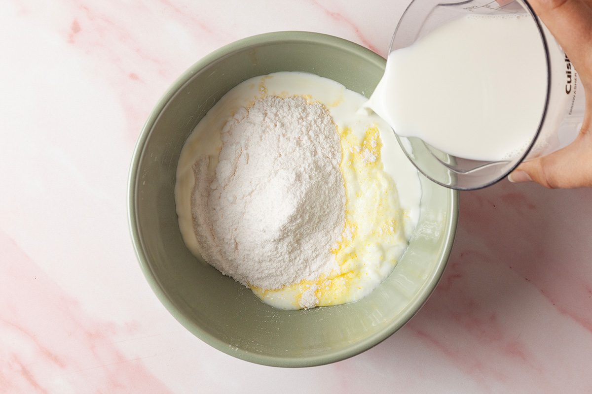 A person pours milk into a mixing bowl filled with flour, sugar, and melted butter on a light pink and white marble surface.