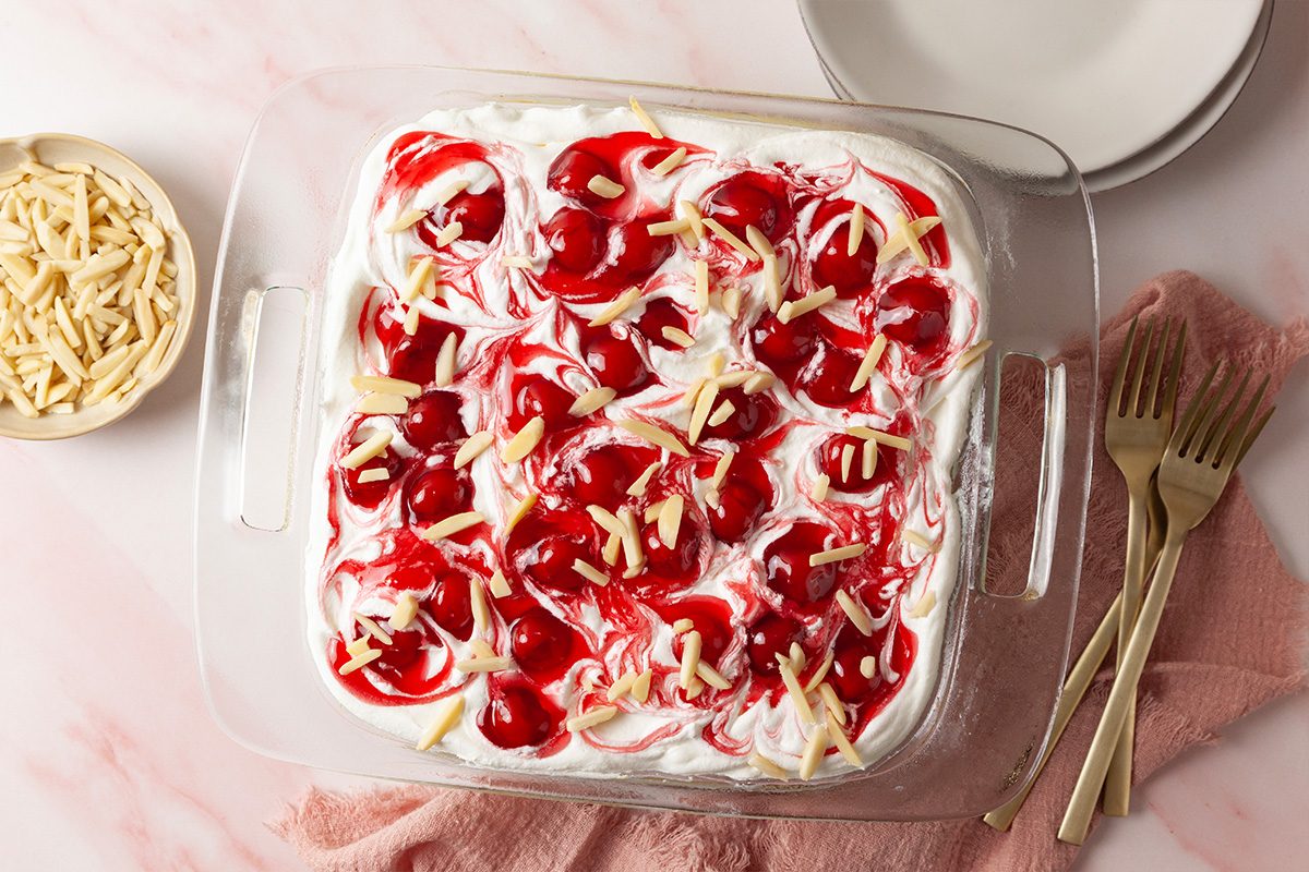 A glass baking dish filled with a dessert topped with whipped cream, cherry pie filling, and slivered almonds sits on a pink surface next to a fork, plate, and a small bowl of slivered almonds.