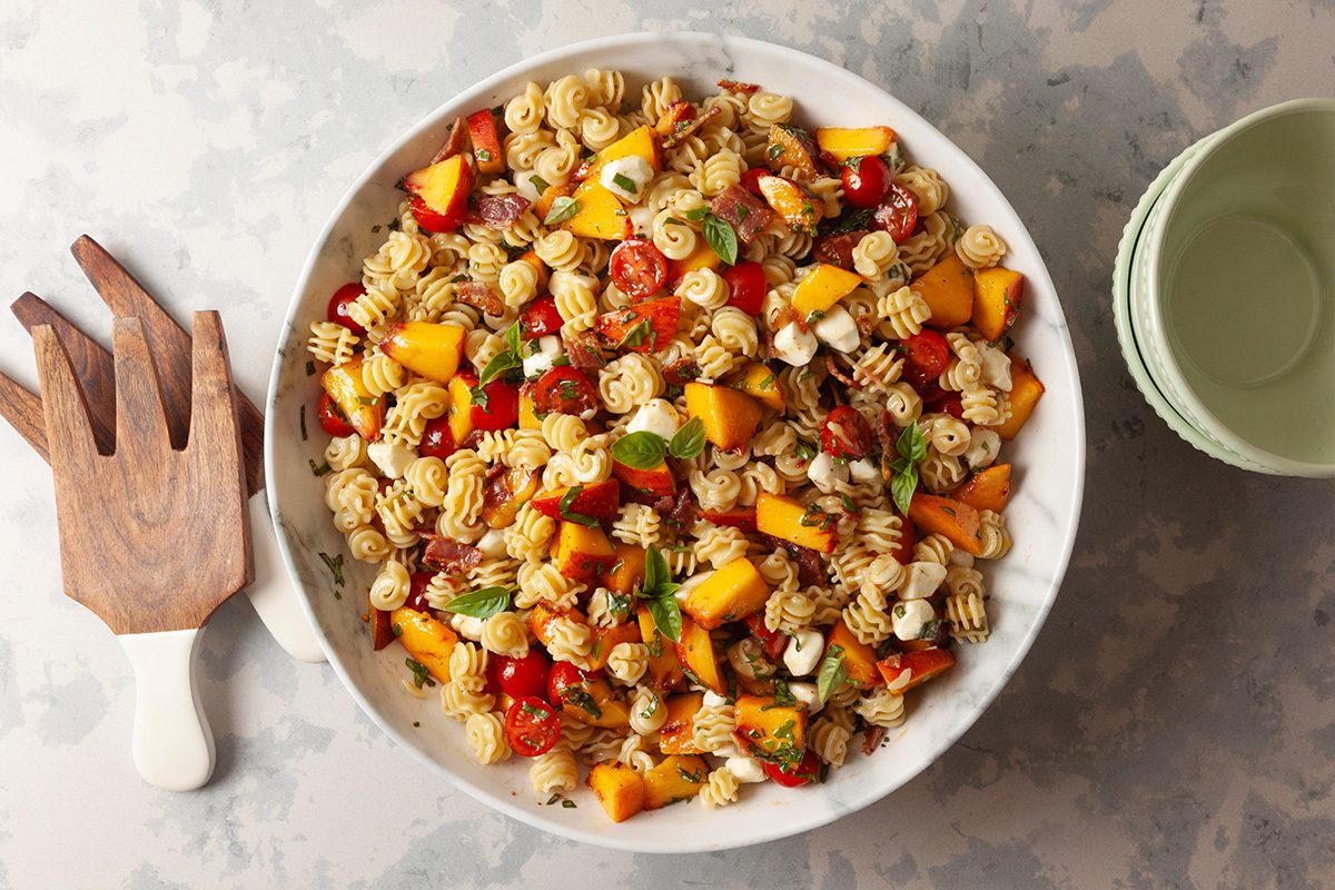 A large white bowl filled with colorful pasta salad, including spiral pasta, cherry tomatoes, mozzarella balls, red bell peppers, and chunks of yellow mango. Wooden salad tongs and empty green bowls sit nearby.