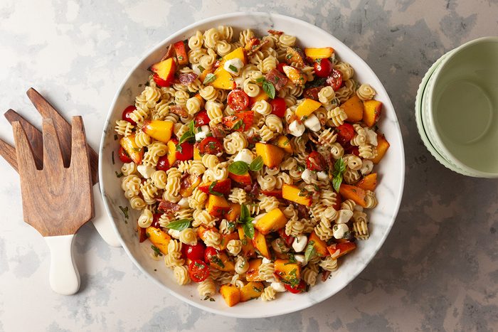 A large white bowl filled with colorful pasta salad, including spiral pasta, cherry tomatoes, mozzarella balls, red bell peppers, and chunks of yellow mango. Wooden salad tongs and empty green bowls sit nearby.