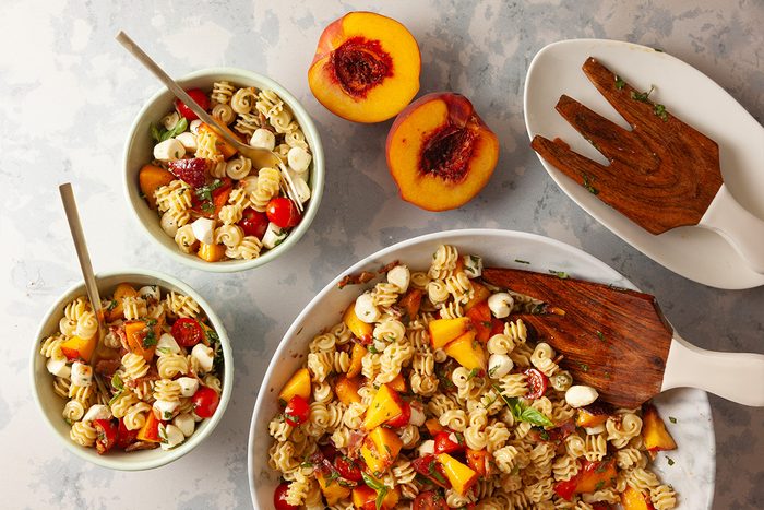 A large bowl and two smaller bowls filled with pasta salad containing cherry tomatoes, mozzarella balls, and diced peaches. Two peach halves and wooden salad utensils are nearby on a light gray surface.