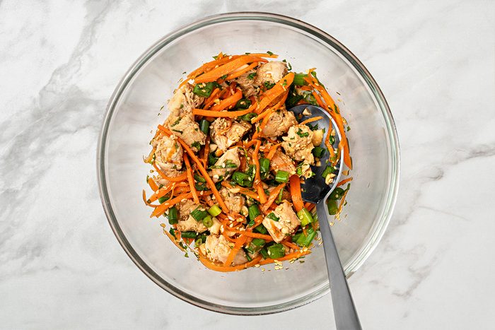 A glass bowl filled with a mixture of cubed tofu, sliced carrots, chopped green onions, and herbs, with a metal spoon resting inside. The bowl is placed on a light marble surface.