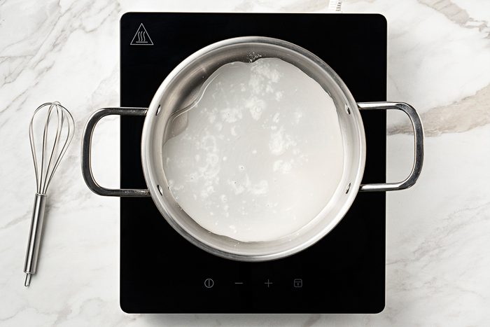 A stainless steel pot with milk is heating on a black induction cooktop, with a metal whisk placed nearby on a white marble countertop.
