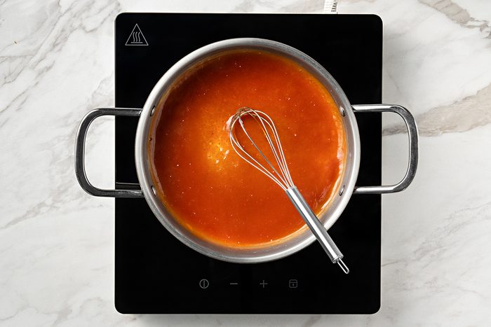 A saucepan filled with orange-red sauce and a metal whisk sits on a black induction cooktop, placed on a white marble countertop.