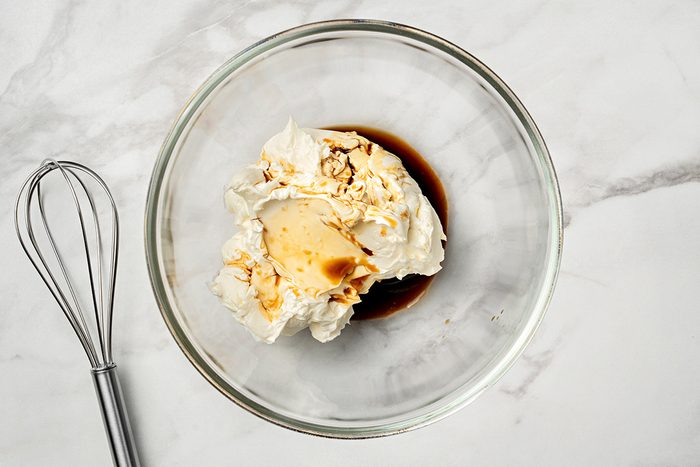 A glass bowl with cream cheese, vanilla extract, and sugar on a white marble surface, next to a metal whisk.