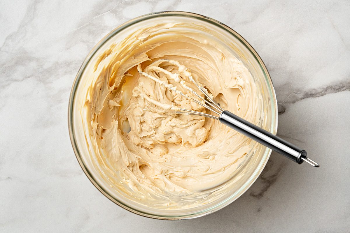 A glass bowl filled with creamy, light beige frosting being mixed with a metal whisk, placed on a white marble surface.