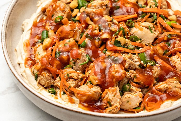 Close-up of a bowl filled with shredded chicken, sliced green onions, carrots, peanuts, and a red sauce on a creamy base. The dish appears to be garnished with sesame seeds and herbs.