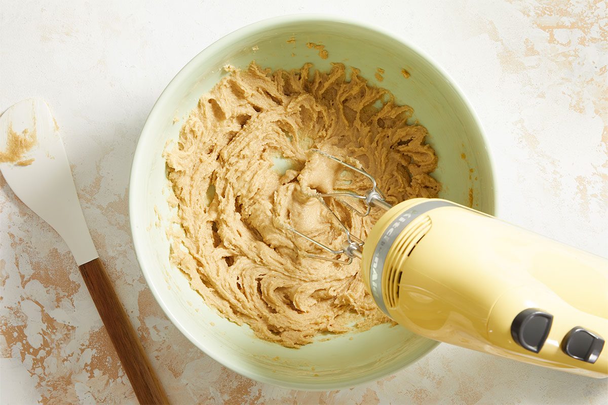 A yellow hand mixer blending light brown cookie dough in a green bowl, with a spatula resting nearby on a white surface.
